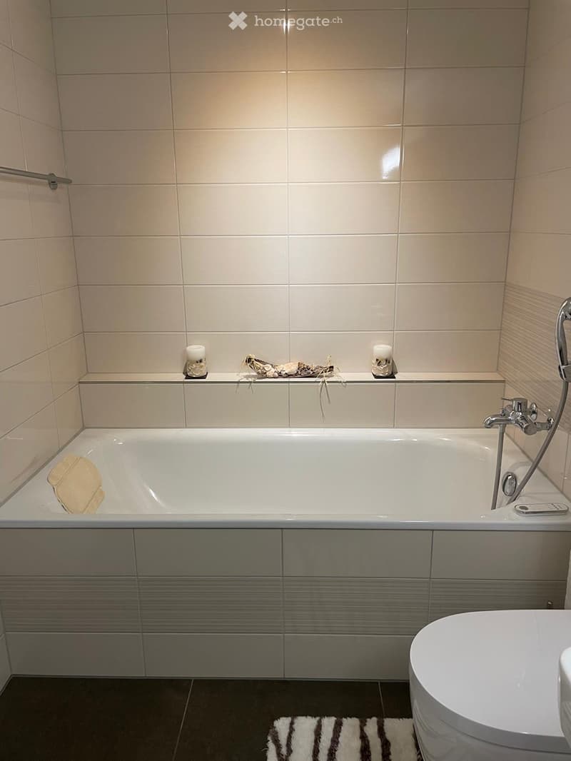 Modern bathroom with a bathtub, sink, and toilet. The bathtub has a modern faucet and a decorative candle on the edge. A small bath mat is placed on the floor near the sink.
