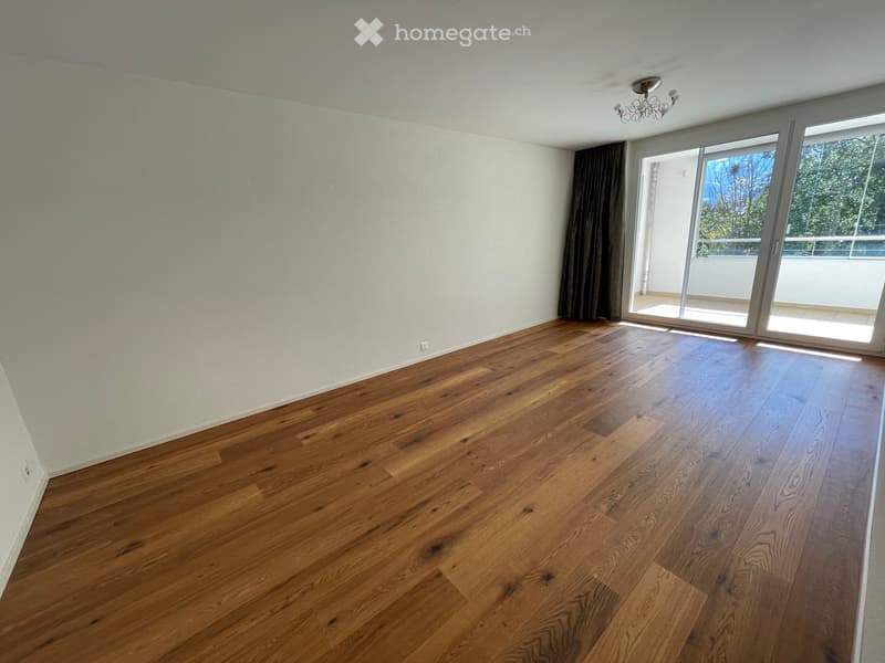 Empty room with wooden flooring, white walls, large sliding glass door leading to a balcony, and a chandelier on the ceiling.