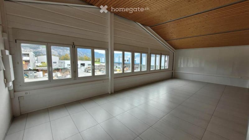 Empty room with large windows offering a view of the surrounding area, including buildings and mountains. The room features a tiled floor and wooden ceiling.