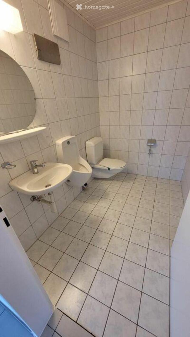 A small, clean bathroom with white tiled walls and floor, featuring a sink, toilet, urinal, and a mirror above the sink.