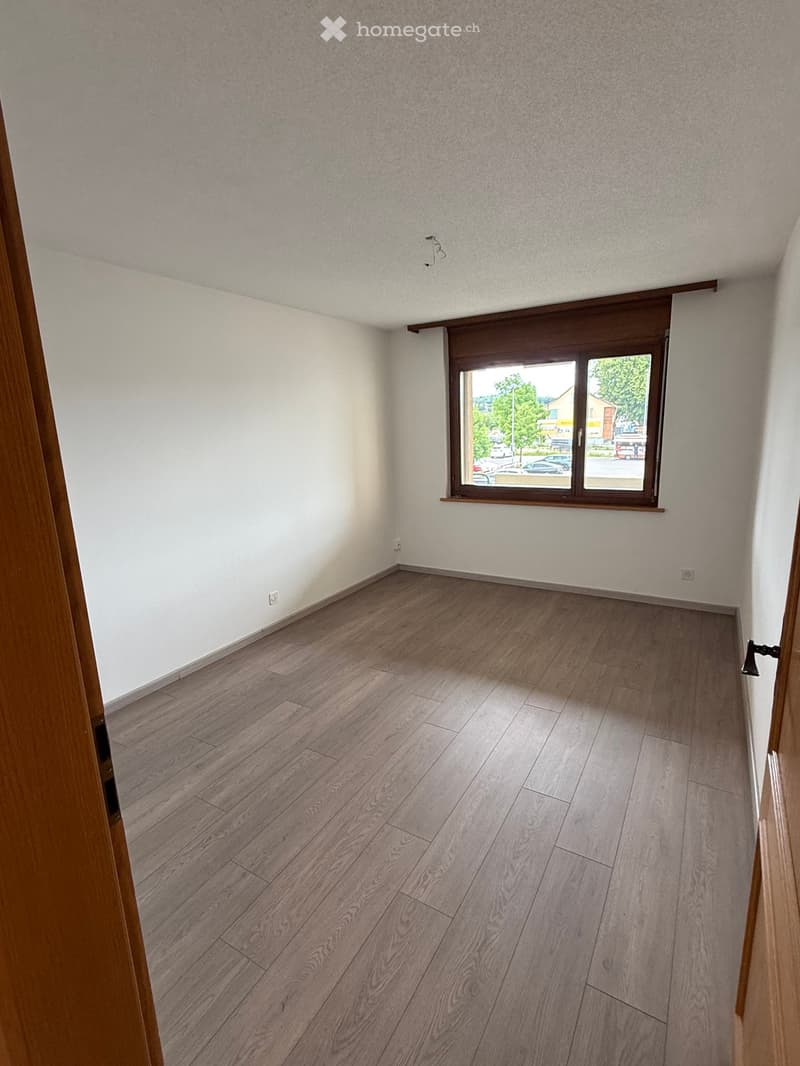 Empty room with light-colored walls, wooden flooring, and a large window with a view of the street.
