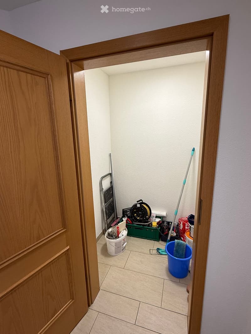 A small utility room with tiled floor, white walls, and various cleaning supplies including buckets, a mop, a ladder, and tools.