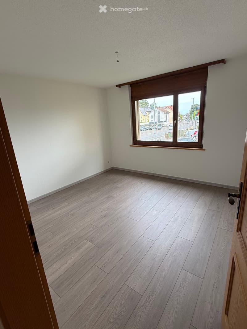 Empty room with light-colored wooden flooring and white walls, featuring a large window with wooden frame and blinds, overlooking a street with parked cars and buildings.
