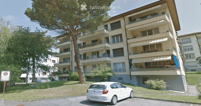 Multi-story residential building with balconies, awnings, and surrounding greenery.