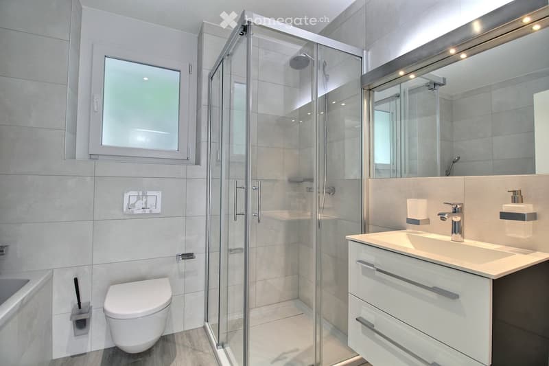 Modern bathroom with a glass-enclosed shower, toilet, sink, and bathtub. Features include a large mirror, sleek fixtures, and a window for natural light.