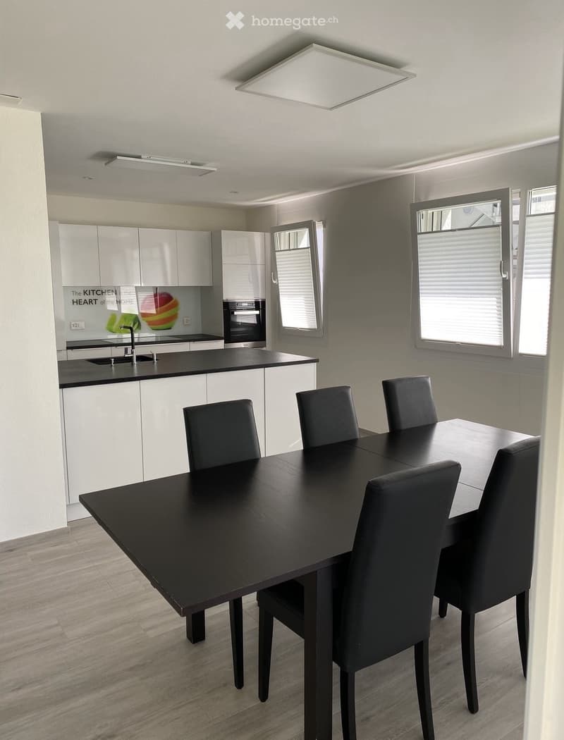 A modern dining room with a large black dining table and chairs, white kitchen cabinets, a kitchen area with an oven and a sink, white tiled flooring, and large windows with white blinds.