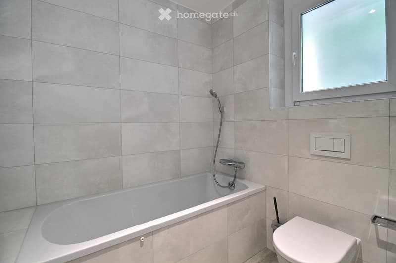 Modern bathroom with a bathtub, toilet, showerhead, and a window with frosted glass. The walls are tiled in light gray, and there is a control panel for the shower on the wall.