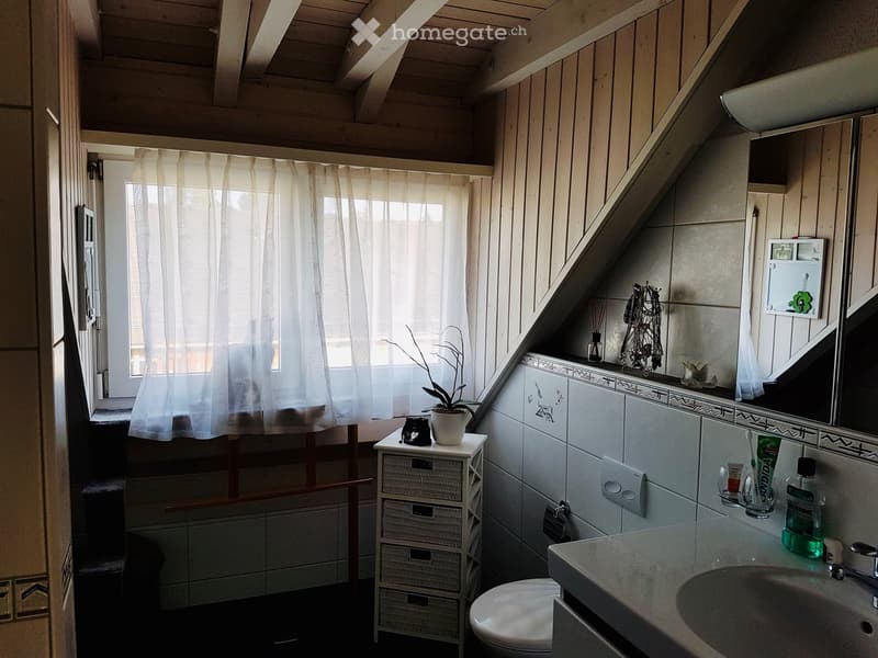 Bathroom with a sink, toilet, mirror, wicker cabinet, and a small table with a plant and other items. The room has wooden paneling and a window with a curtain.