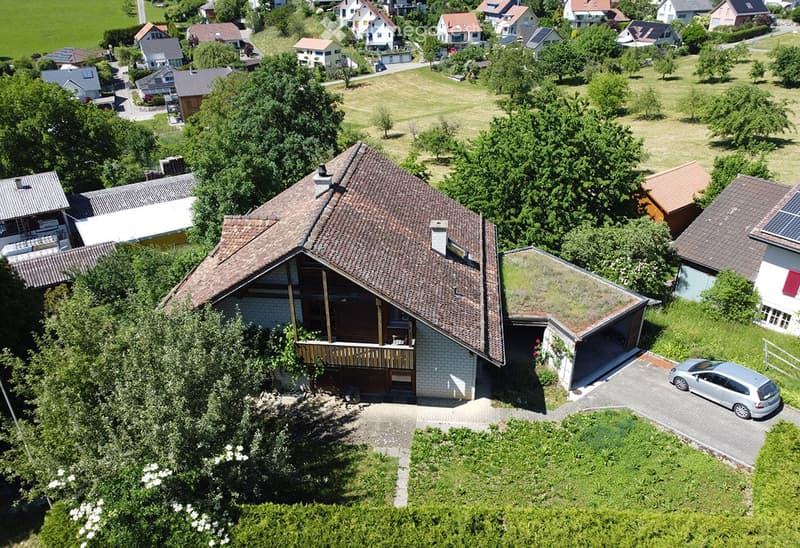 A two-story house with a brown tiled roof, a balcony, and a parking area with a car. The house is surrounded by greenery and trees, with neighboring houses visible in the background.