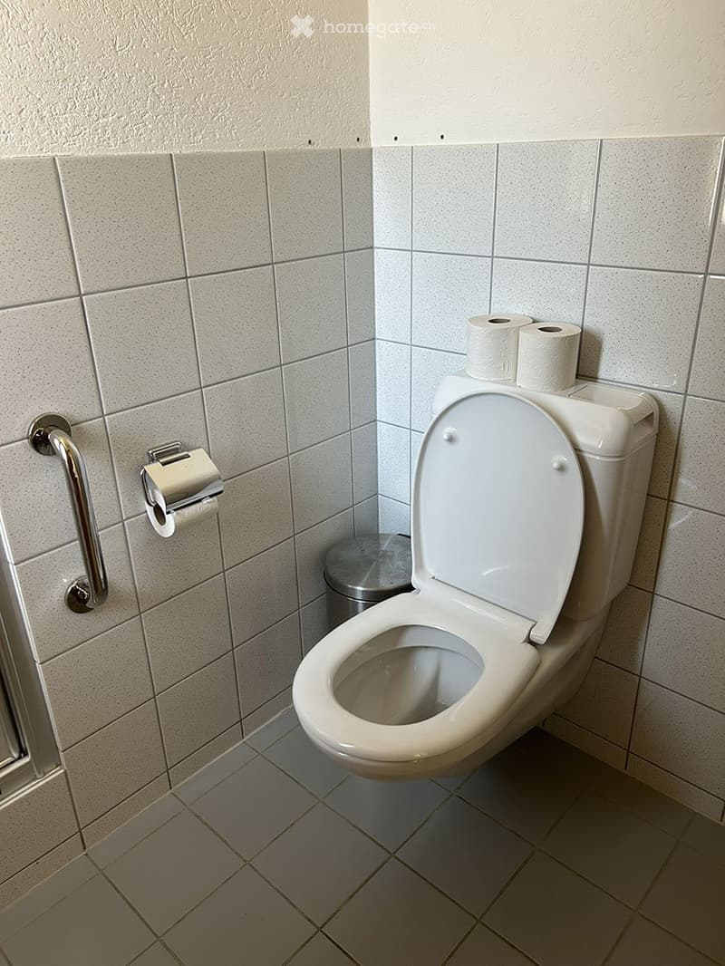 Clean bathroom with white tiled walls and floor, toilet with lid up, toilet paper rolls on holder and shelf, and a small trash can.