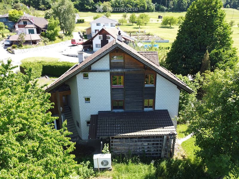 A two-story house with a tiled roof, white brick exterior, and wooden accents. The house features multiple windows and a small attached structure, surrounded by greenery and trees.
