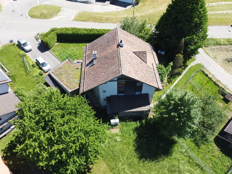 A two-story house with a tiled roof, surrounded by a well-maintained lawn and trees. The house features a garage and a driveway with parked cars.