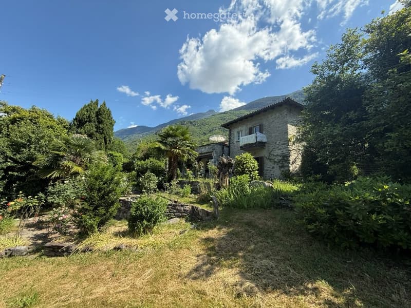 A rustic stone house surrounded by a lush garden with various plants, trees, and a scenic mountain view in the background.