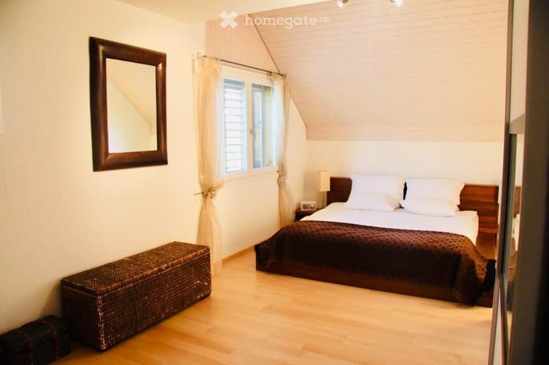 Cozy bedroom with wooden flooring, a large bed with white pillows and a dark brown duvet, a wicker storage box, a nightstand with a lamp, and a large mirror on the wall.