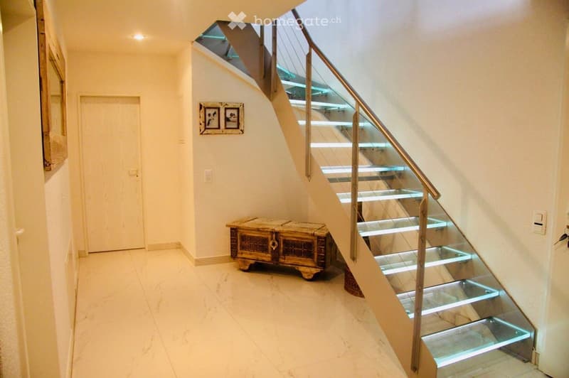 Modern hallway with a sleek staircase featuring glass steps and a metal railing, illuminated steps, a wooden chest, and a framed picture on the wall.