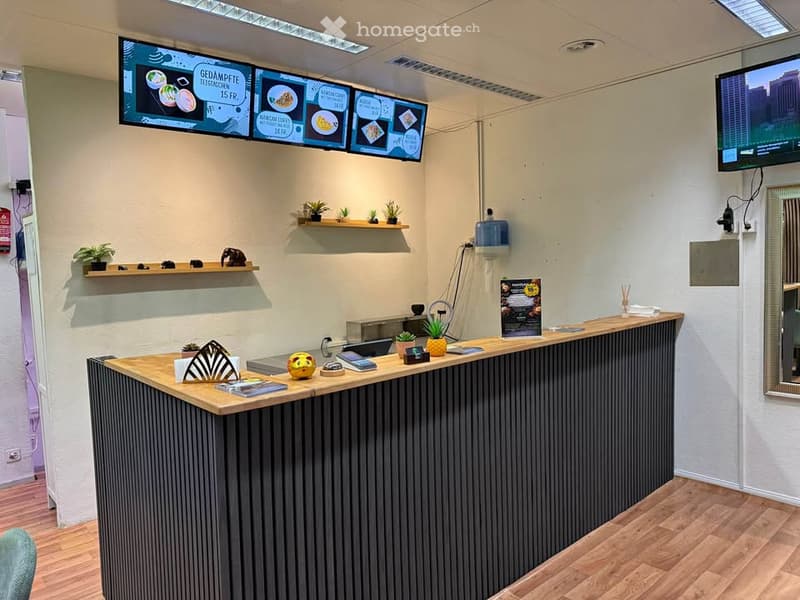 Reception area with a modern wooden desk, digital menu screens, small potted plants, and a mirror on the wall.