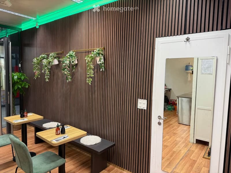 A modern, stylish room with wooden flooring, dark vertical wall panels, and a mix of seating and tables. Potted plants are displayed on the wall and in corners, and a small kitchen area is visible through an open doorway.