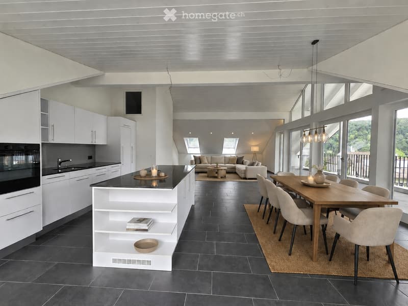Open-plan kitchen and living area with white cabinets, black countertops, a central island, a dining table with chairs, and a sofa in the background. The space features large windows and a modern design.