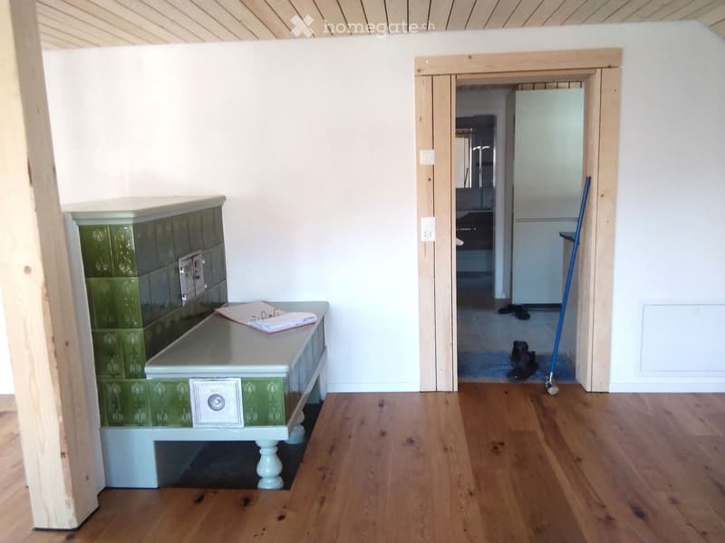 A small, well-lit hallway with wooden flooring, a green-tiled bench, and a table with papers on it. A doorway leads to another room with visible furniture.