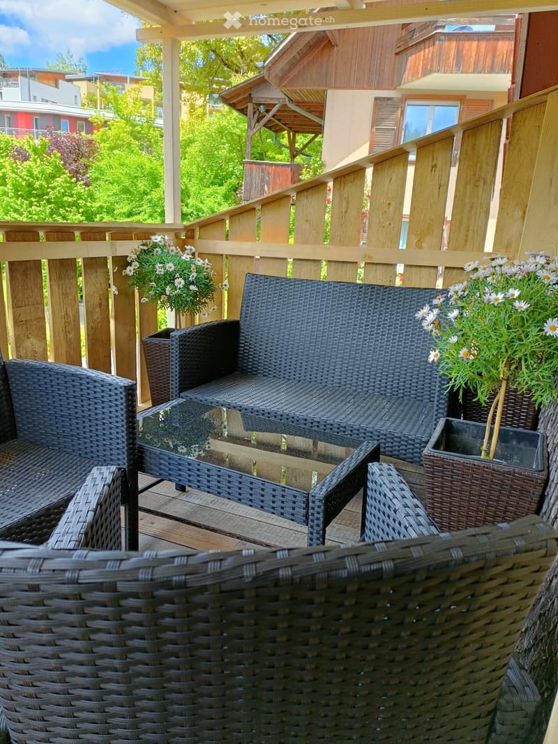 A wooden balcony with wicker furniture, including a sofa, chairs, and a small table, decorated with white flower pots.