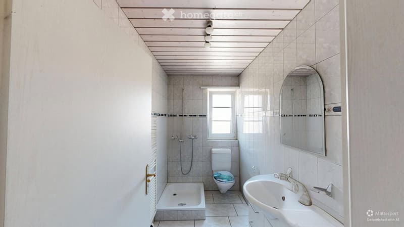 Bathroom with white tiles, a toilet, a sink, a shower, and a mirror. The room has a window and is well-lit.