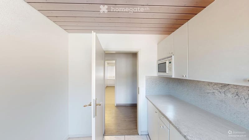 A modern kitchen with white cabinets, a microwave, and a marble countertop. The hallway features wooden flooring and white walls.