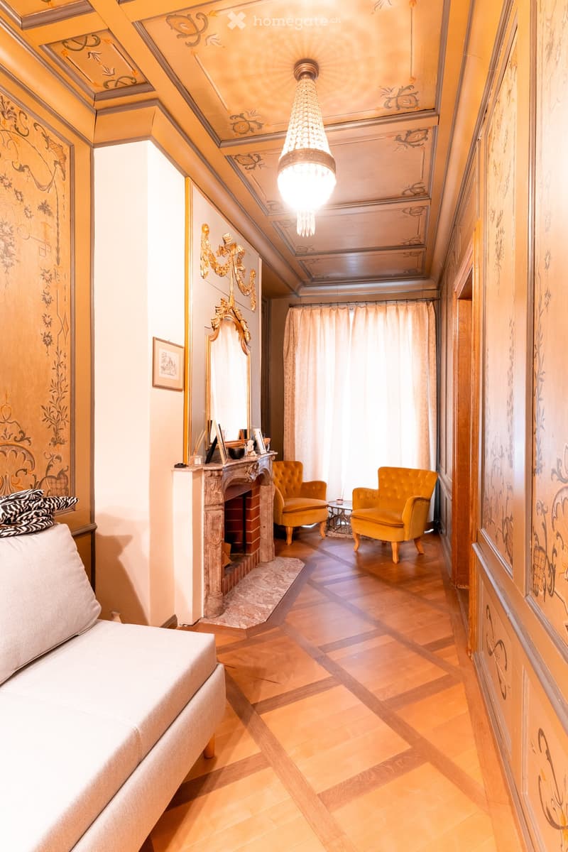 Elegant room with a fireplace, two armchairs, a sofa, a chandelier, and a mirror. The room features ornate wooden walls and a slanted ceiling.