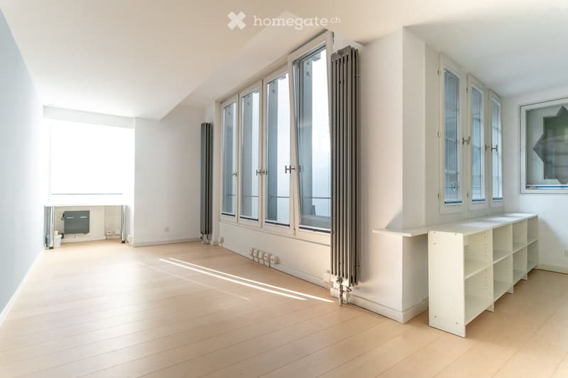 Bright, empty room with wooden flooring, large windows, built-in shelving unit, and radiator. No furniture or personal items present.