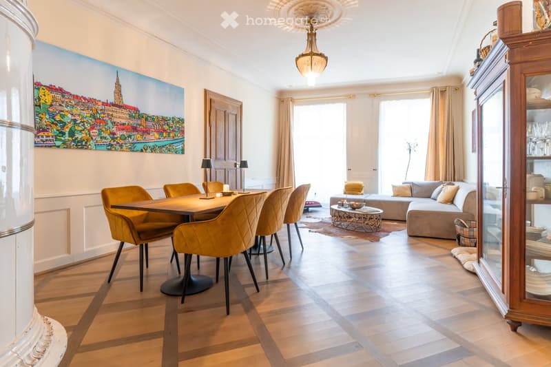 Open-plan living and dining area with wooden flooring, a large sofa, a coffee table, a dining table with chairs, and a cabinet with glass doors.