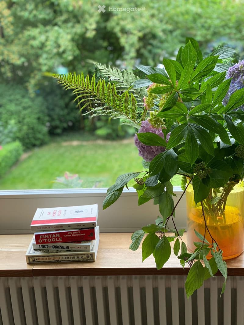 A wooden table with a vase of flowers and a stack of books, placed near a window with a view of a green garden.