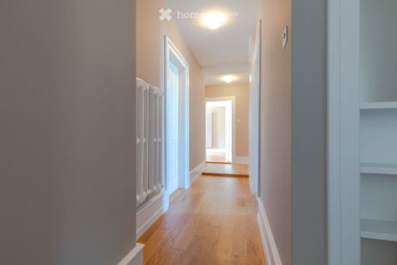 A long, narrow hallway with wooden flooring, beige walls, and white baseboards. The hallway features a radiator on the left wall and a thermostat on the right wall. The ceiling has recessed lighting, and the hallway leads to an open doorway at the end.