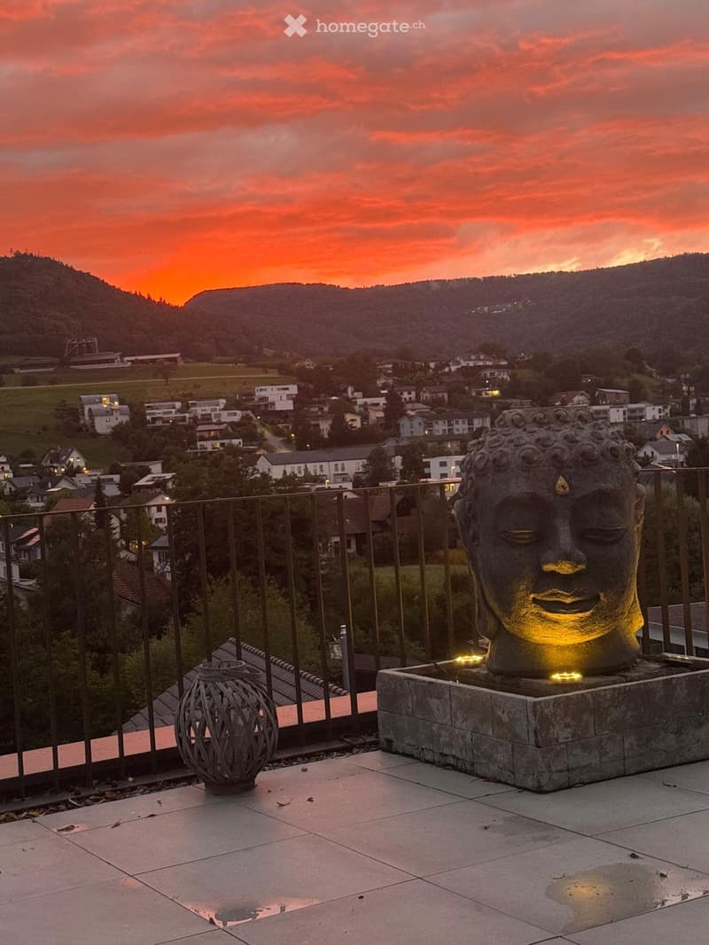 Outdoor area with tiled floor, metal railing, Buddha statue, small lantern, sunset backdrop with mountains and village.