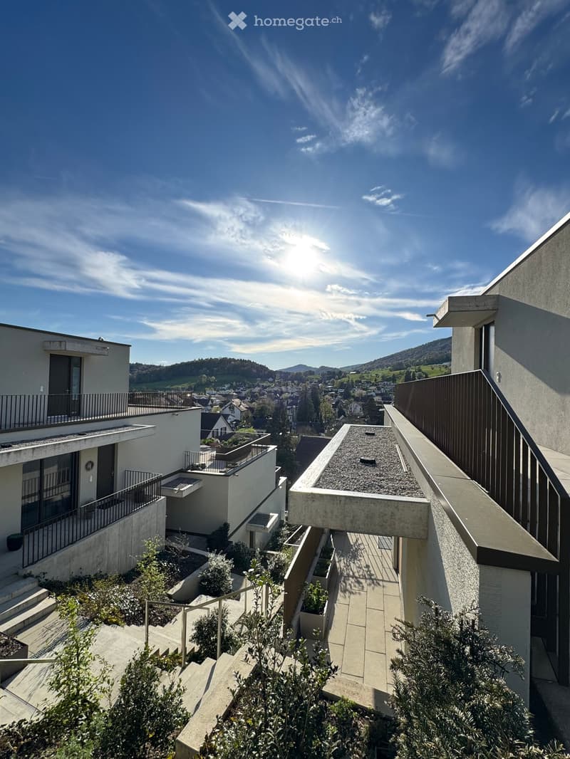 Modern building with multiple balconies, concrete and glass design, surrounded by greenery, elevated view of the town and mountains