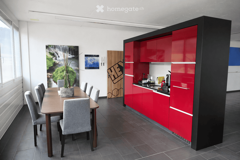 Modern dining area with a wooden table, six grey chairs, and a red kitchen cabinet unit. The kitchen features a coffee machine, sink, and faucet. A vase with dried plants sits on the table, and a large waterfall painting adorns the wall.