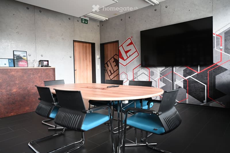 Modern conference room with a round wooden table surrounded by black and blue chairs, a large flat-screen screen on the wall, and a wooden cabinet with picture frames on top.