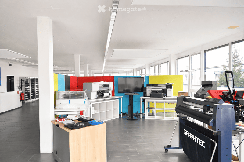 Modern office space with large windows, multiple printers, a screen, tables, and shelves.