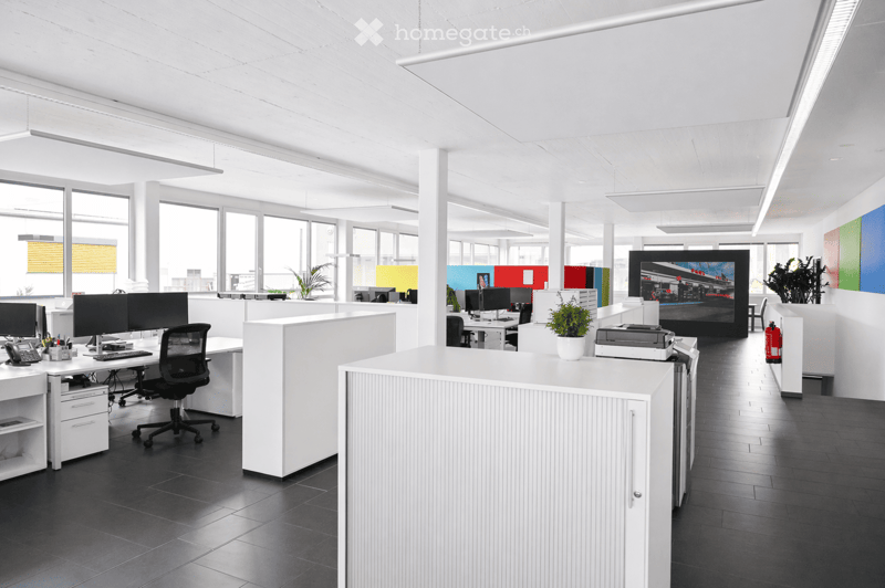Modern office space with white desks, black chairs, computer monitors, and a large screen displaying an image. The room features large windows with blinds, potted plants, and a fire extinguisher on the floor.