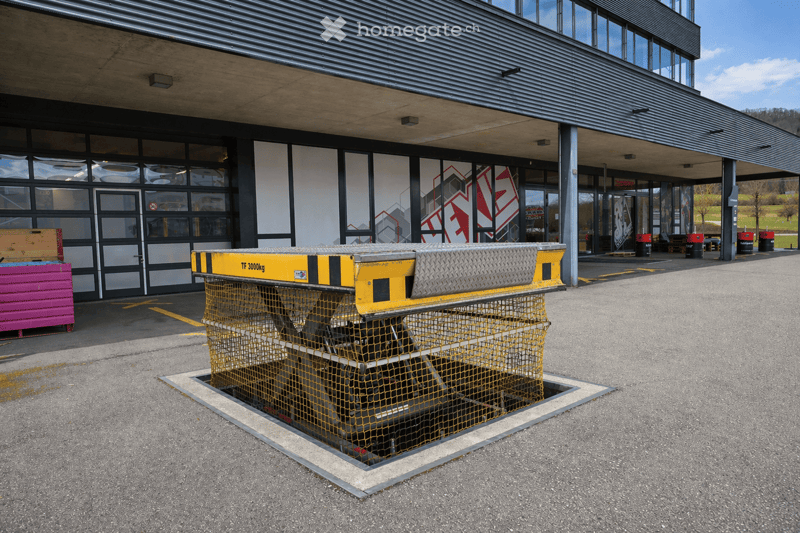 Modern building entrance with large glass windows and doors, featuring a raised ground floor with a covered walkway. A yellow loading platform with a protective net is visible in the foreground.