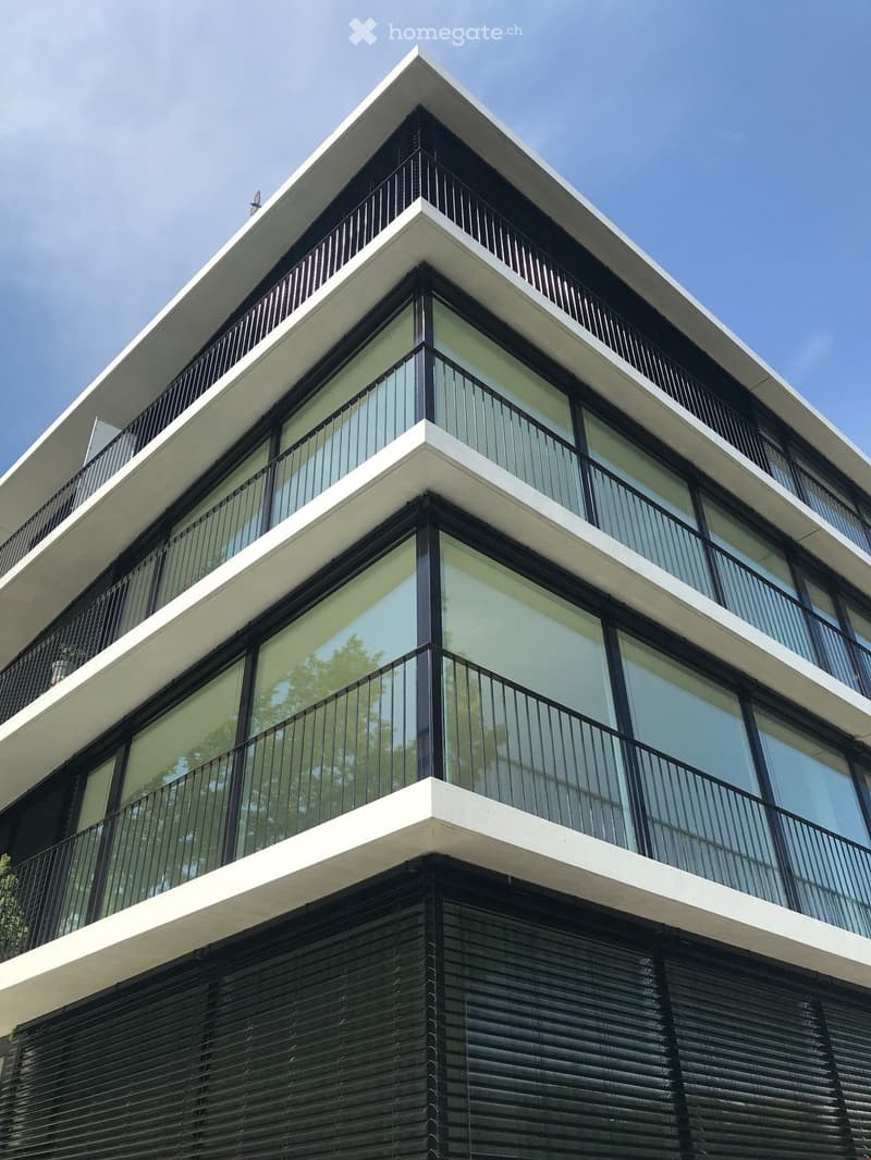Modern multi-story building with large glass windows and black metal railings on balconies. The structure features a clean, contemporary design with white and black accents.