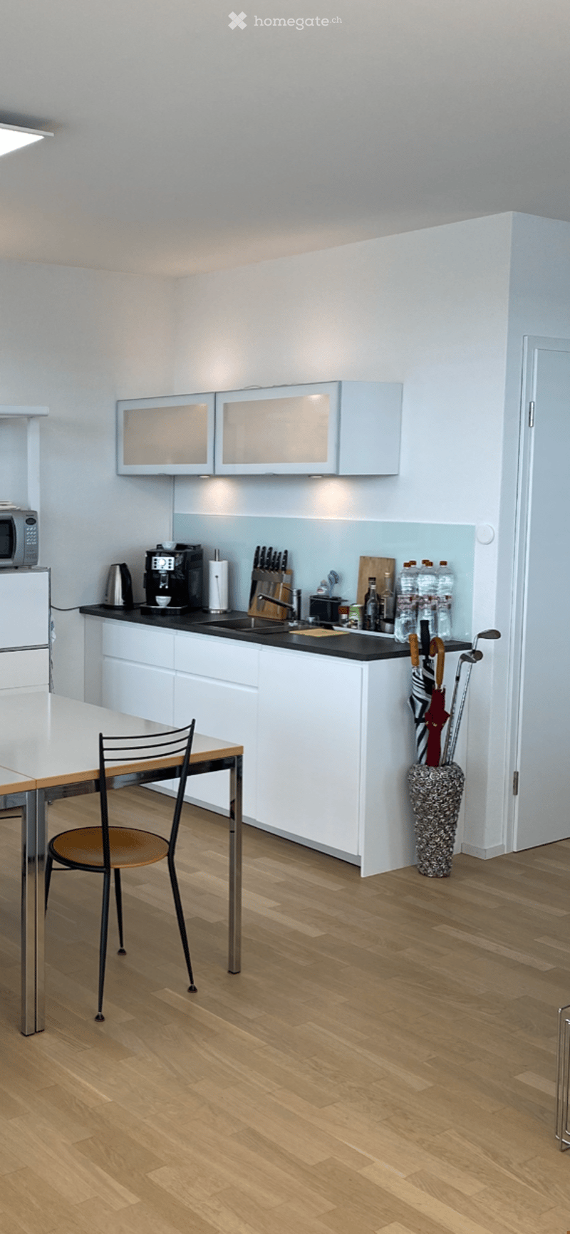 Modern kitchen with white cabinets, black countertops, stainless steel appliances, and a small dining area with a table and chair. Wooden flooring and a decorative umbrella stand are also visible.