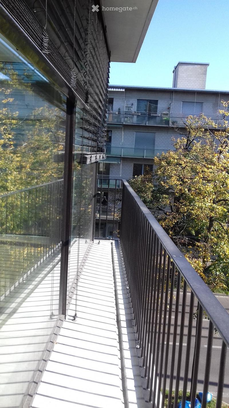 Modern balcony with glass railing, metal balustrade, and steps leading to a rooftop area. The building in the background has a contemporary design with large windows and a balcony.
