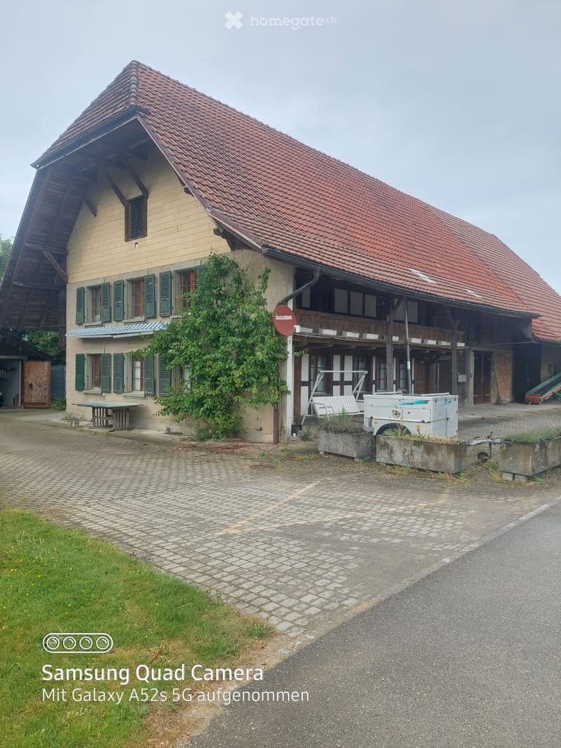 House. Two-story house with red-tiled roof, green shutters, and a covered porch area. The building has a mix of wooden and plaster exterior walls, with a small garden and paved driveway in front.