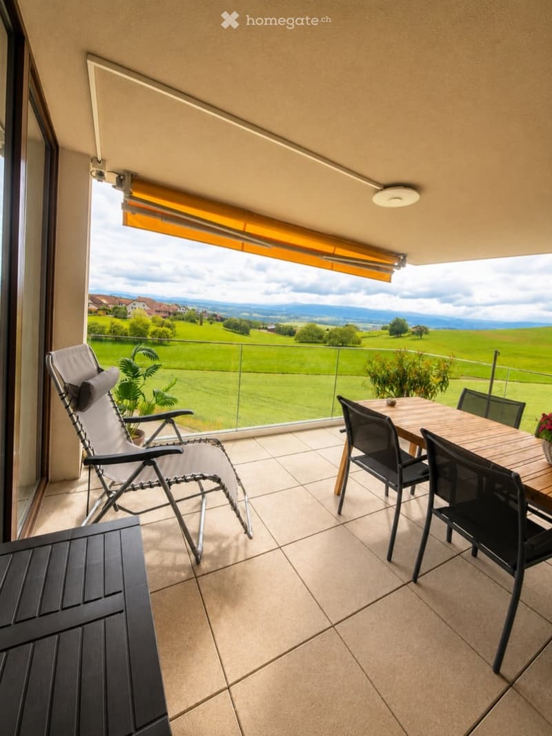Balcony with tiled floor, glass railing, and scenic countryside view. Includes a wooden table with four black chairs, a folding lounge chair, and potted plants.