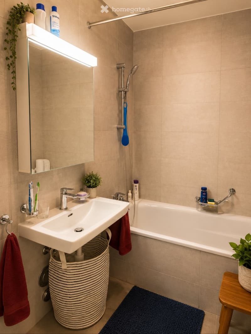 Modern bathroom with a white sink, bathtub, and shower area. Features include a toothbrush holder, a small plant, a towel, and a mirror with built-in lighting.