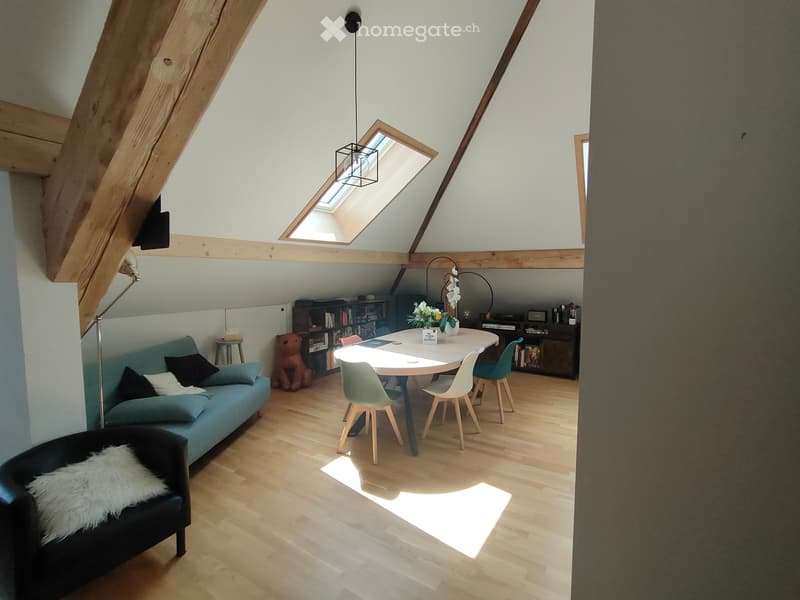 Bright, open loft-style room with wooden floors, slanted ceilings, and skylight. Features a blue sofa, black armchair, dining table with chairs, bookshelf, and decorative items.