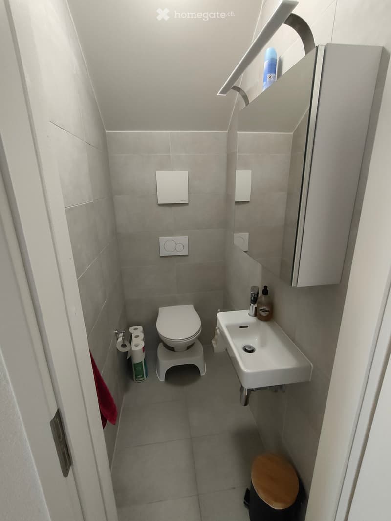 Compact bathroom with a toilet, sink, mirror, and toilet paper rolls. Includes a small wooden stool and a bottle on the sink.
