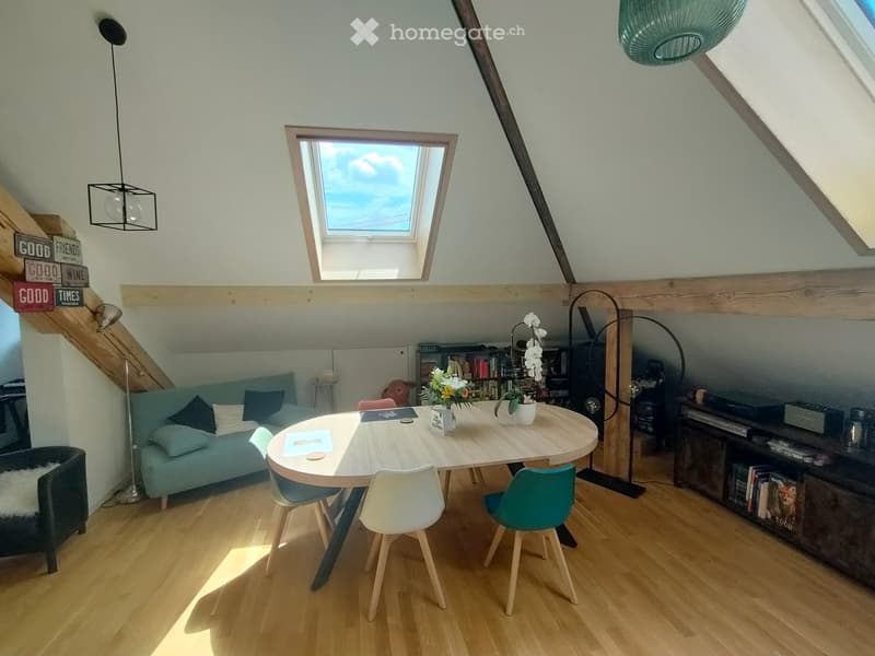 Bright, airy room with wooden floors, a round wooden table with four chairs, a light blue sofa, a bookshelf, and a hanging lamp.