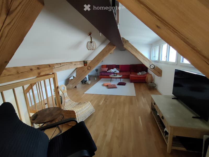 Living room with wooden floors, exposed wooden beams, a red sofa, a coffee table, a wicker chair, and a television on a stand.