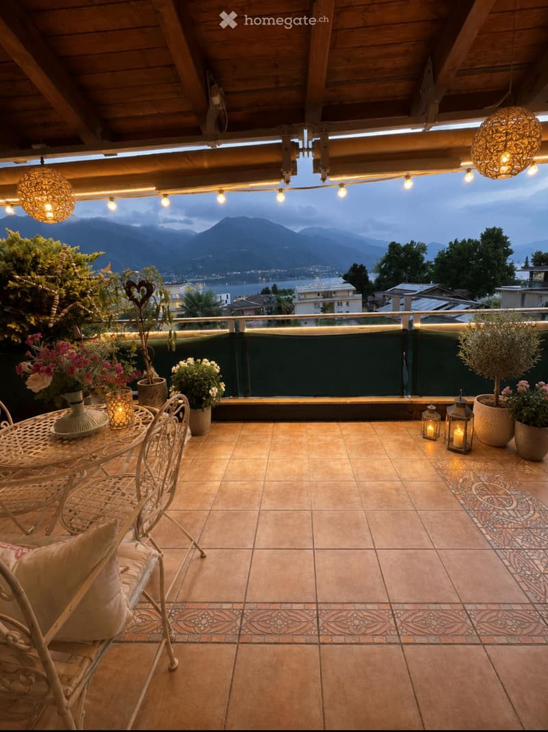 Balcony with terracotta tiles, wooden ceiling, string lights, and a scenic mountain view. Includes a small table, chairs, vases, lanterns, and potted plants.