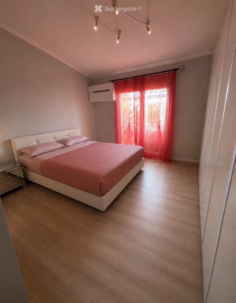 Bedroom with a double bed, pink bedding, nightstand, and large windows with pink curtains. Wooden flooring and modern lighting fixtures.